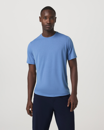 Man wearing a Vuori - Current Tech Tee in Isle Blue and dark pants on a light gray background
