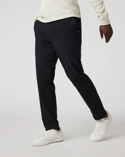 A person wearing black Vuori - Fleet Chino Pant Athletic Slim 30" pants and a white top, standing against a white background.