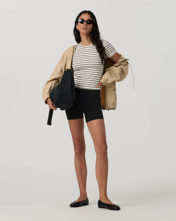 Woman wearing a Vuori - Pose Fitted Tee Ink Stripe black shorts, and sunglasses, holding a beige jacket and black bag on a white background.
