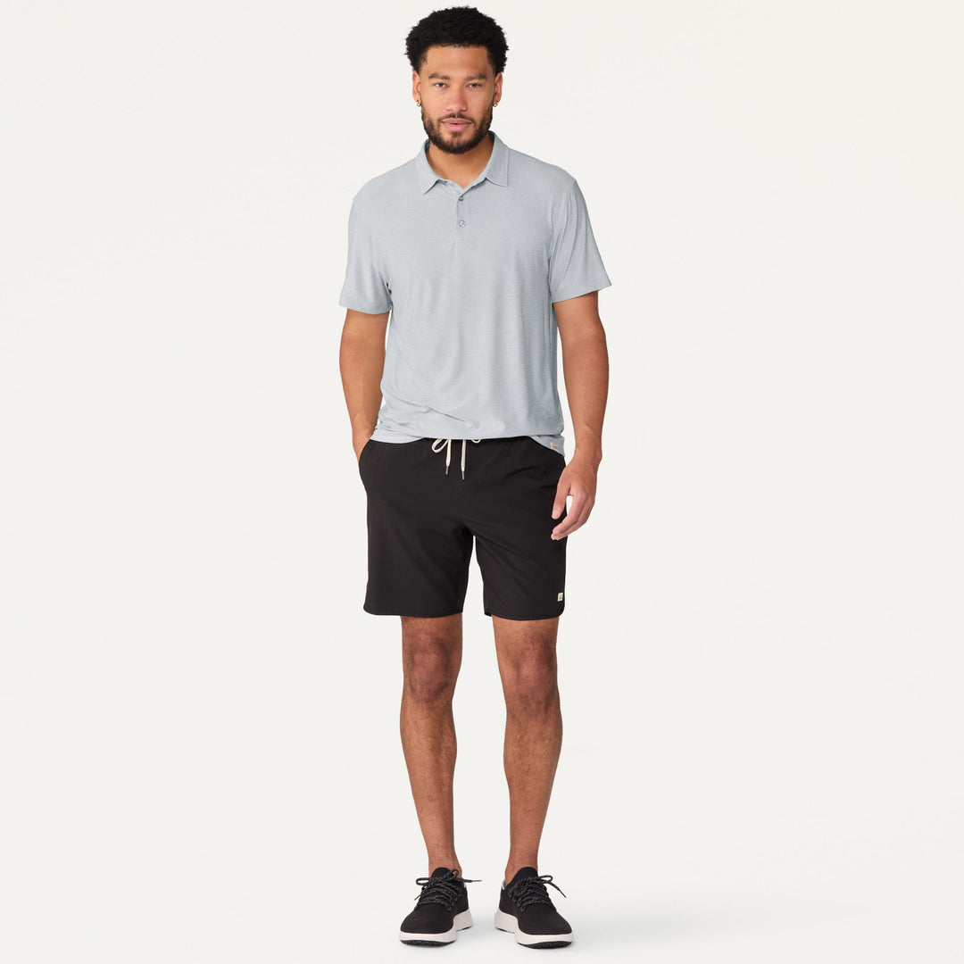 Man wearing a Vuori - Strato Tech Polo in Harbor Grey and black shorts on a white background