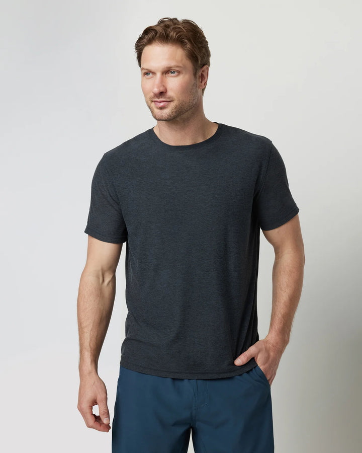 A man wearing a Vuori - Strato Tech Tee in Navy Heather and white pants standing against a white background.