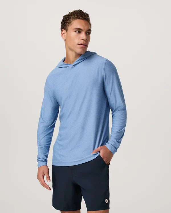 Man wearing a blue Vuori long-sleeve Strato hoodie and black shorts on a light gray background